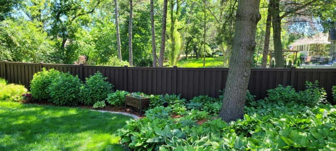 A well-manicured backyard with a wooden fence, lush greenery, and tall trees under a bright sky.