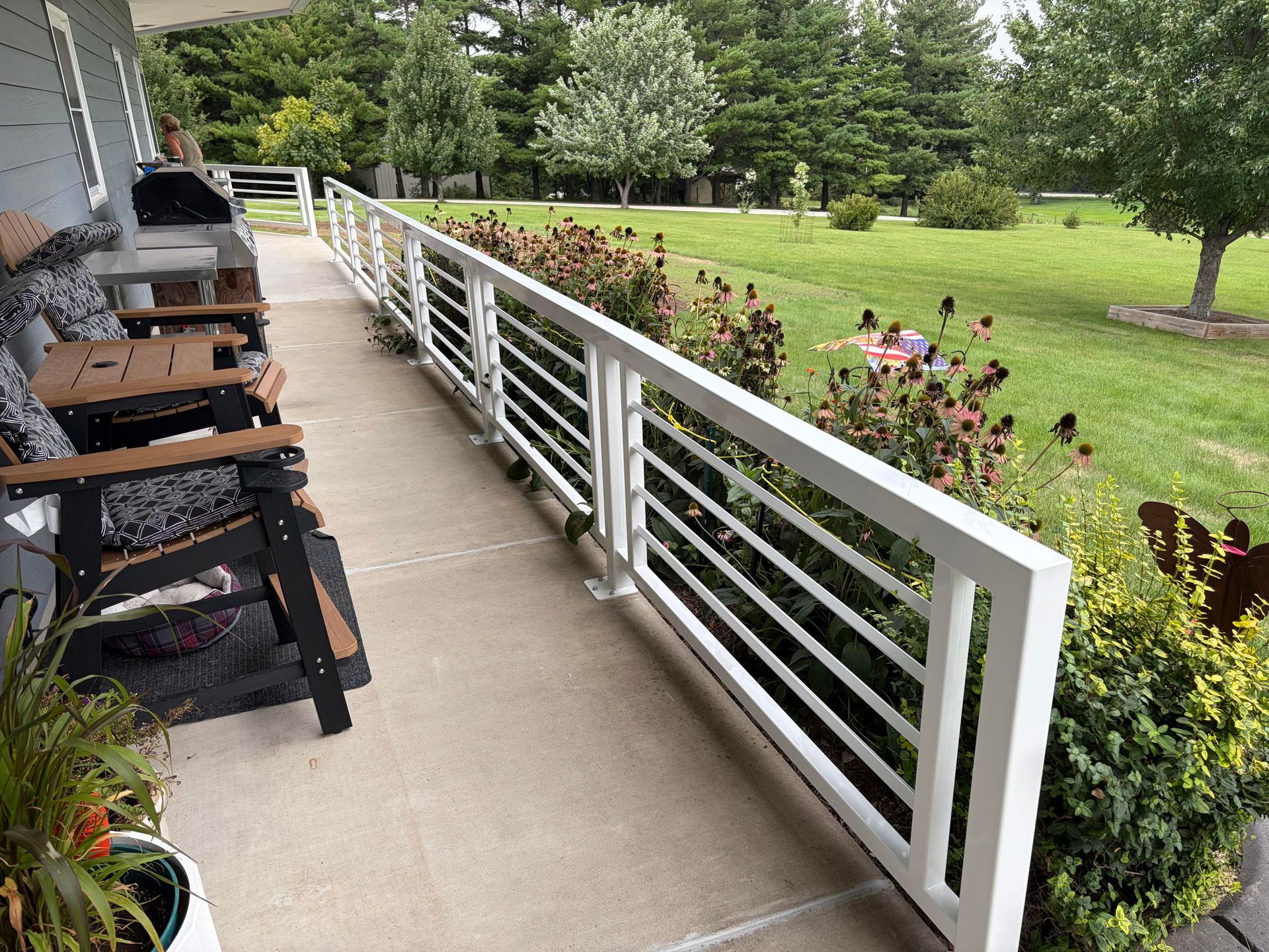 Covered porch with black chairs beside a white railing, overlooking a green lawn and flower bed.