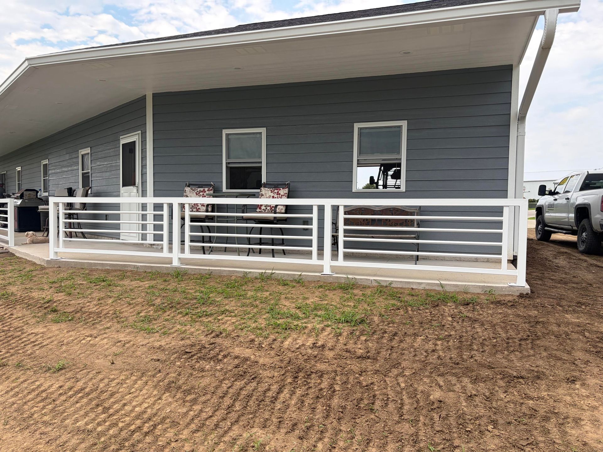 Single-story gray house with white railings and a covered porch on a dirt lot
