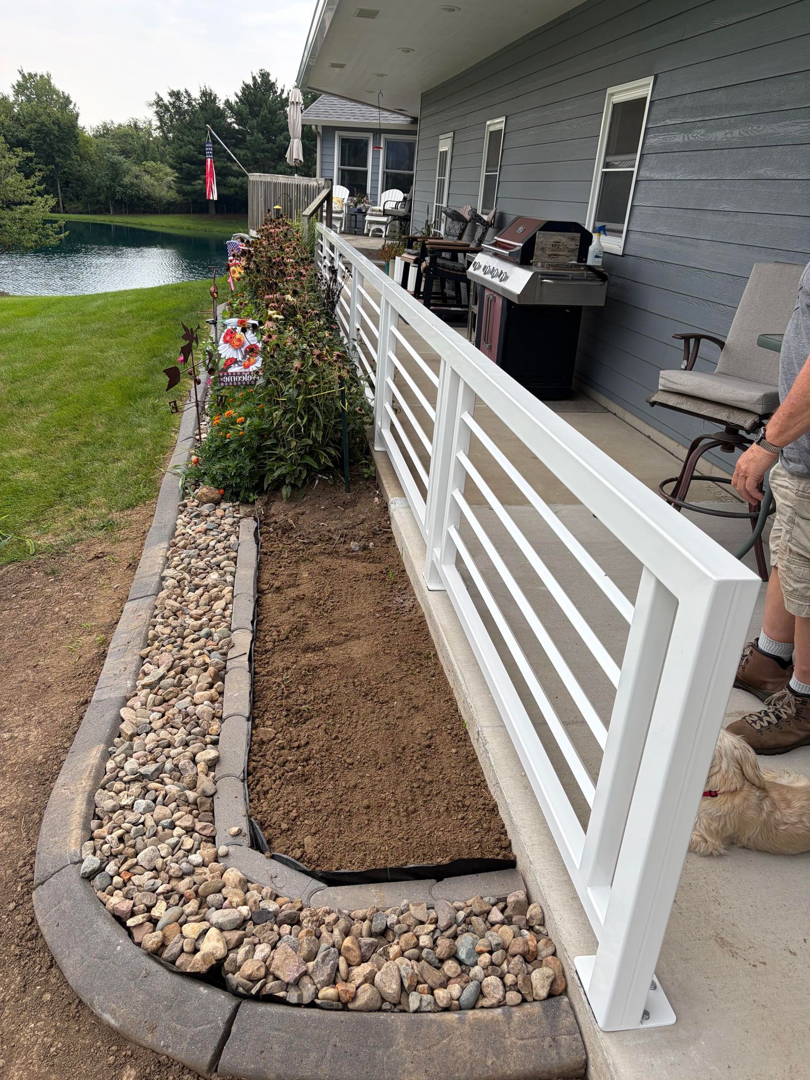 White fence beside a mulched garden bed along a patio, with flowers and a pond in the background