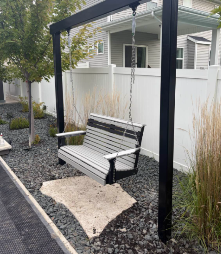 Black porch swing on a gravel patio beside a white fence and gray house, hanging from a dark frame.