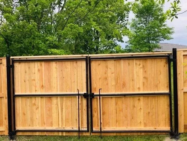 Wooden double gate with black metal frame and latch, in front of a green tree backdrop.