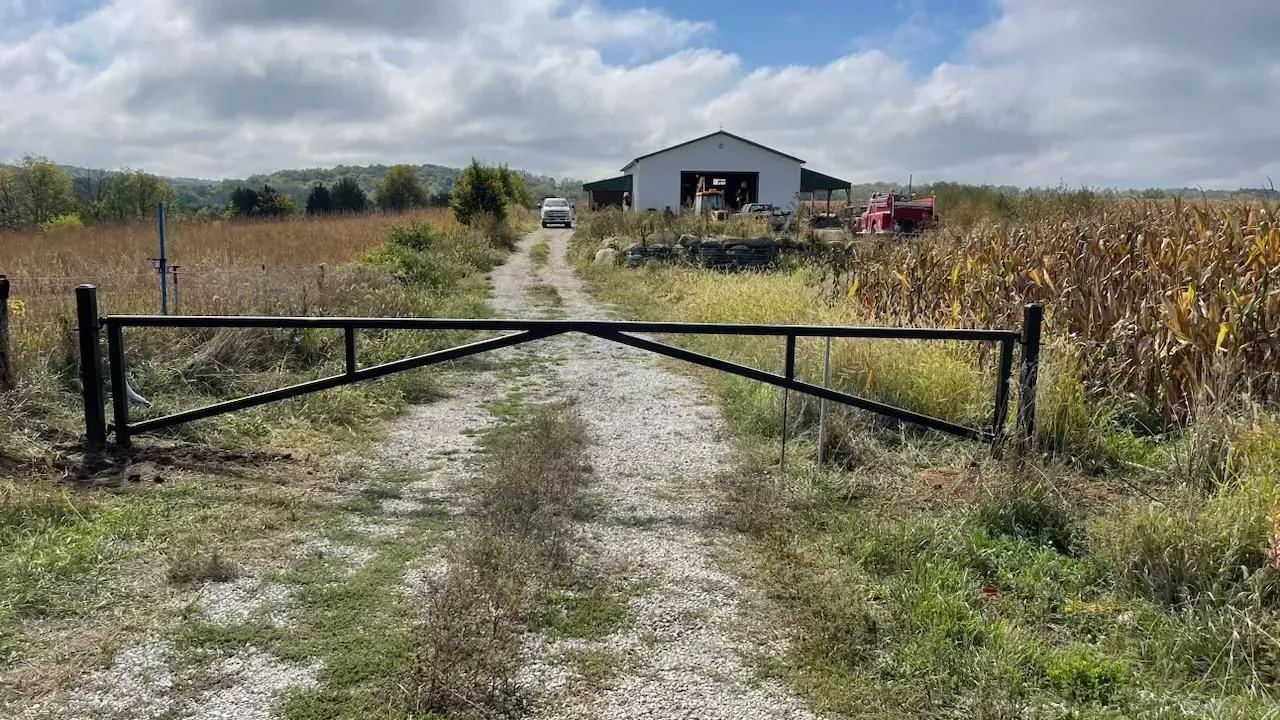 Black gate on a dirt road leading to a white barn in a field under a cloudy sky.