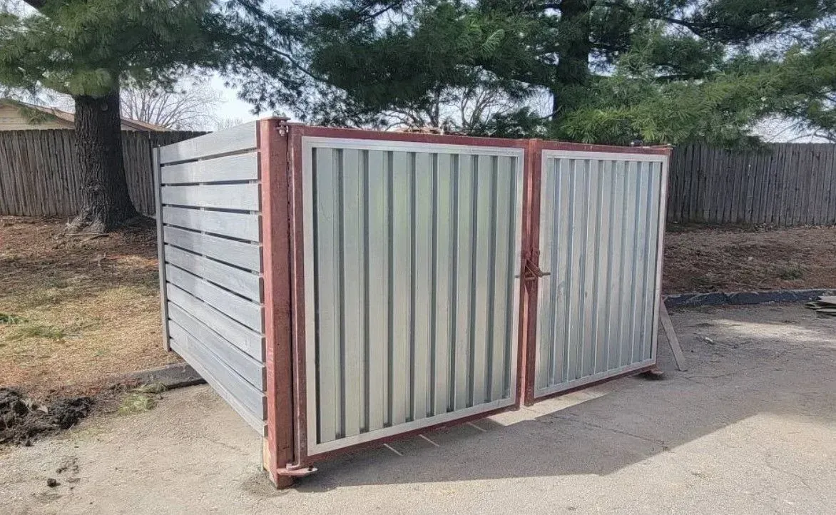 Metal and wood enclosure with two doors, likely for trash cans, outdoors.