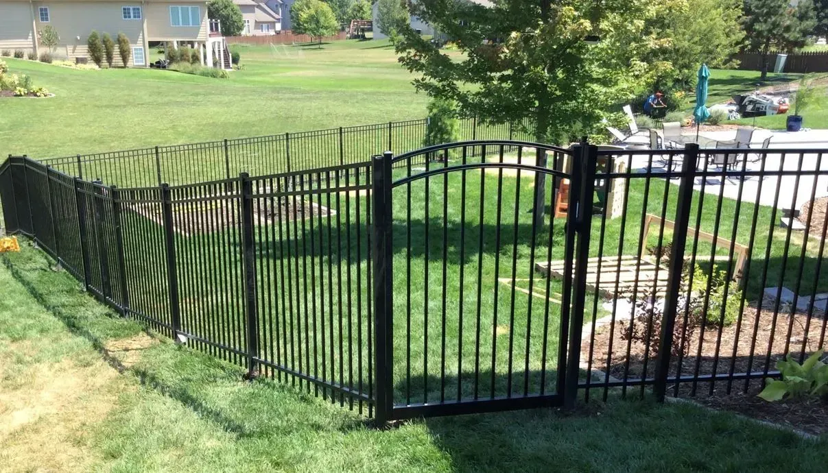 Black metal fence surrounding a grassy backyard with a gate, trees, and a pool area in the distance.