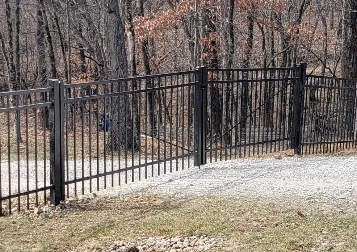 Black metal fence with gate, set against a wooded backdrop and gravel ground.