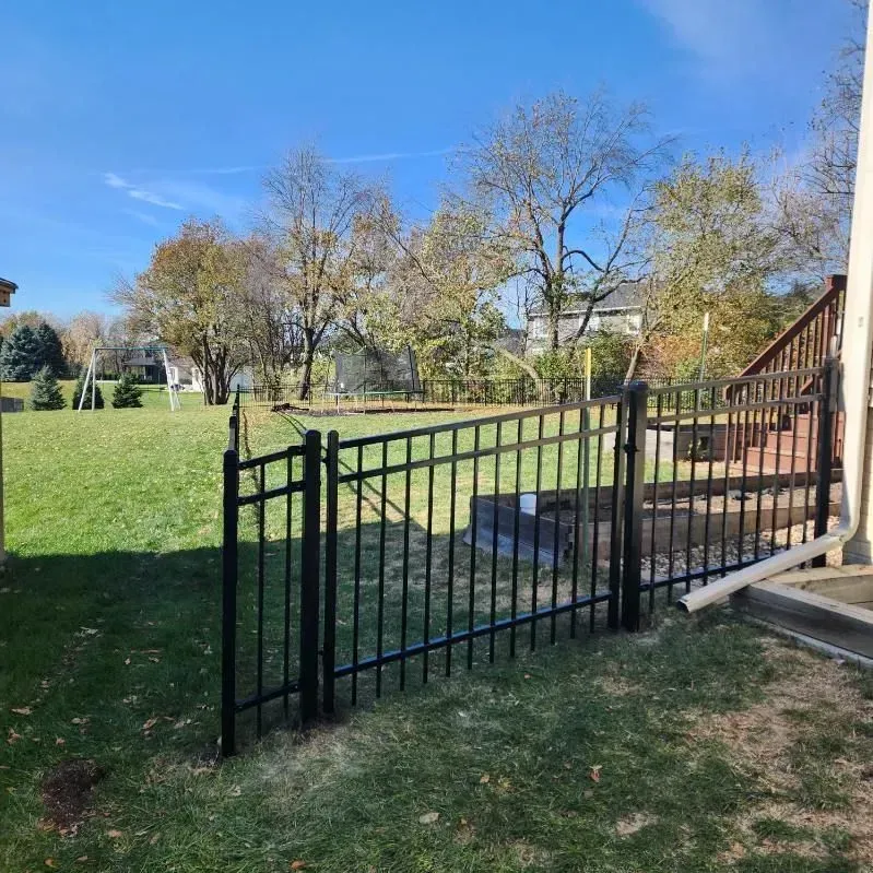 Black metal fence in a yard with a gate, trees, and a house under a blue sky.