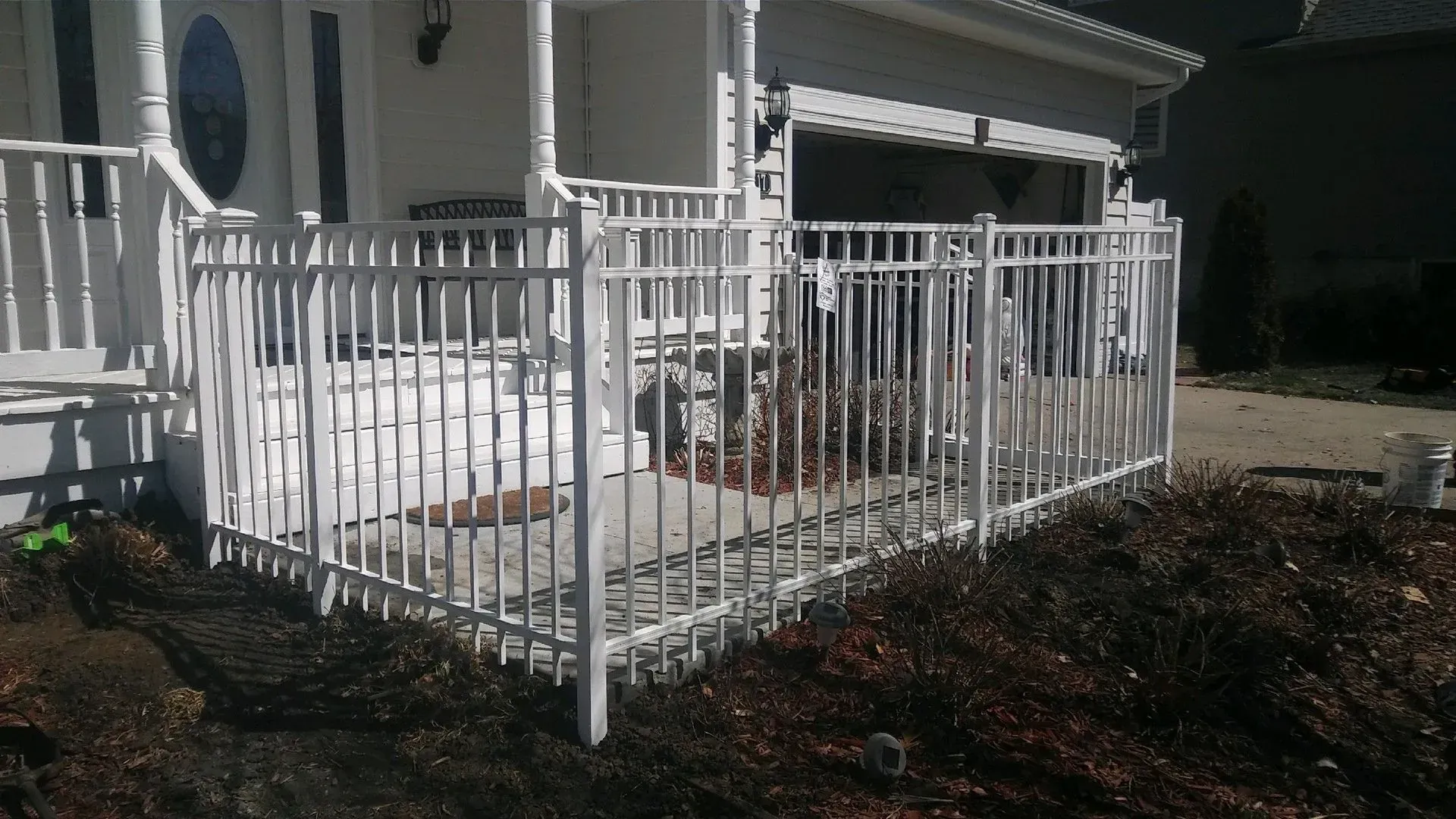 White metal fence encloses a small concrete porch area near a house with a garage.