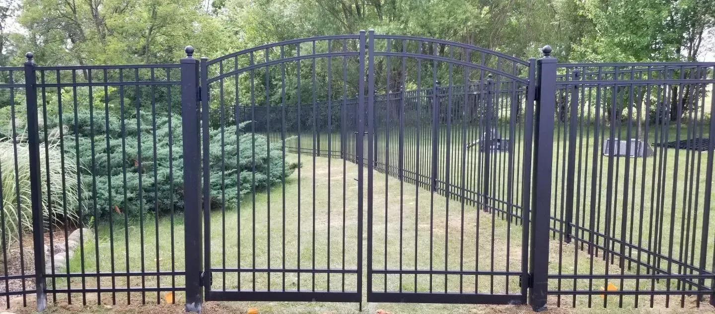 Black metal gate with a matching fence in a grassy yard. Trees in the background.