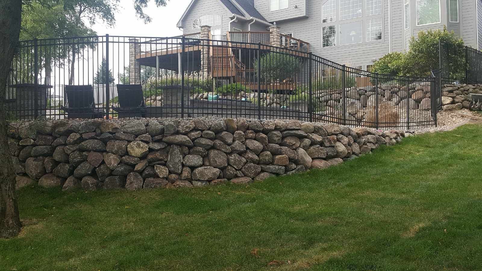Stone retaining wall with black metal fence, overlooking a deck and house.