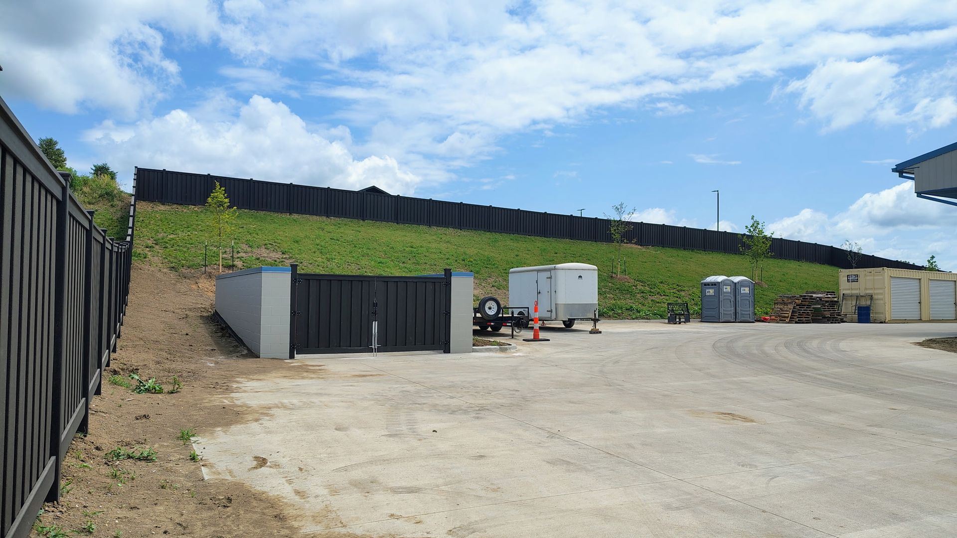 Empty gravel lot beside black fencing and green hillside under a bright sky
