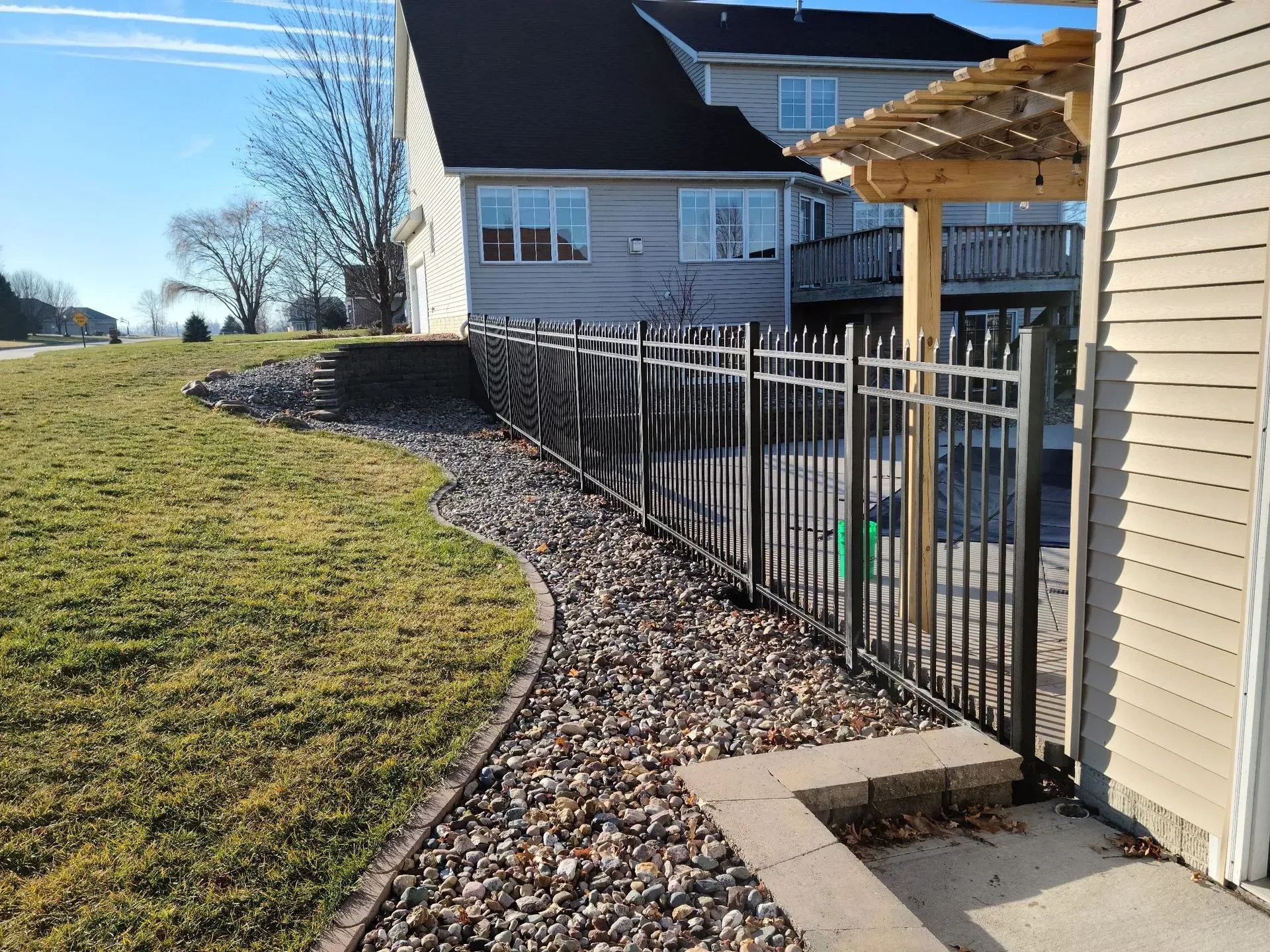 Black metal fence along a gravel bed beside a building, lawn, and retaining wall.