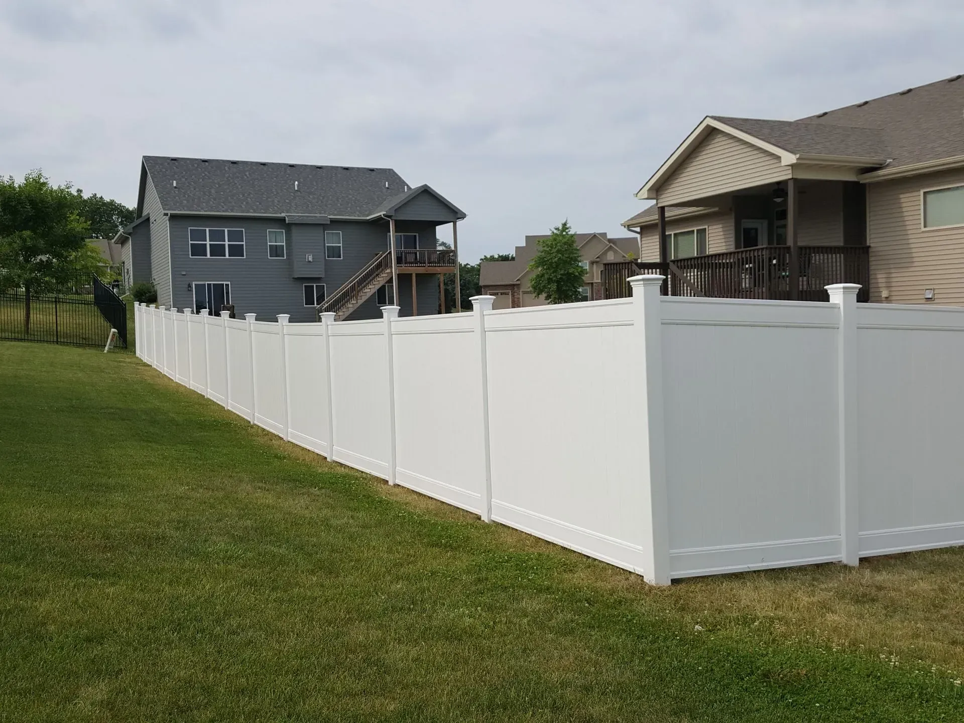 White vinyl fence in a grassy backyard, separating two suburban houses under a cloudy sky.