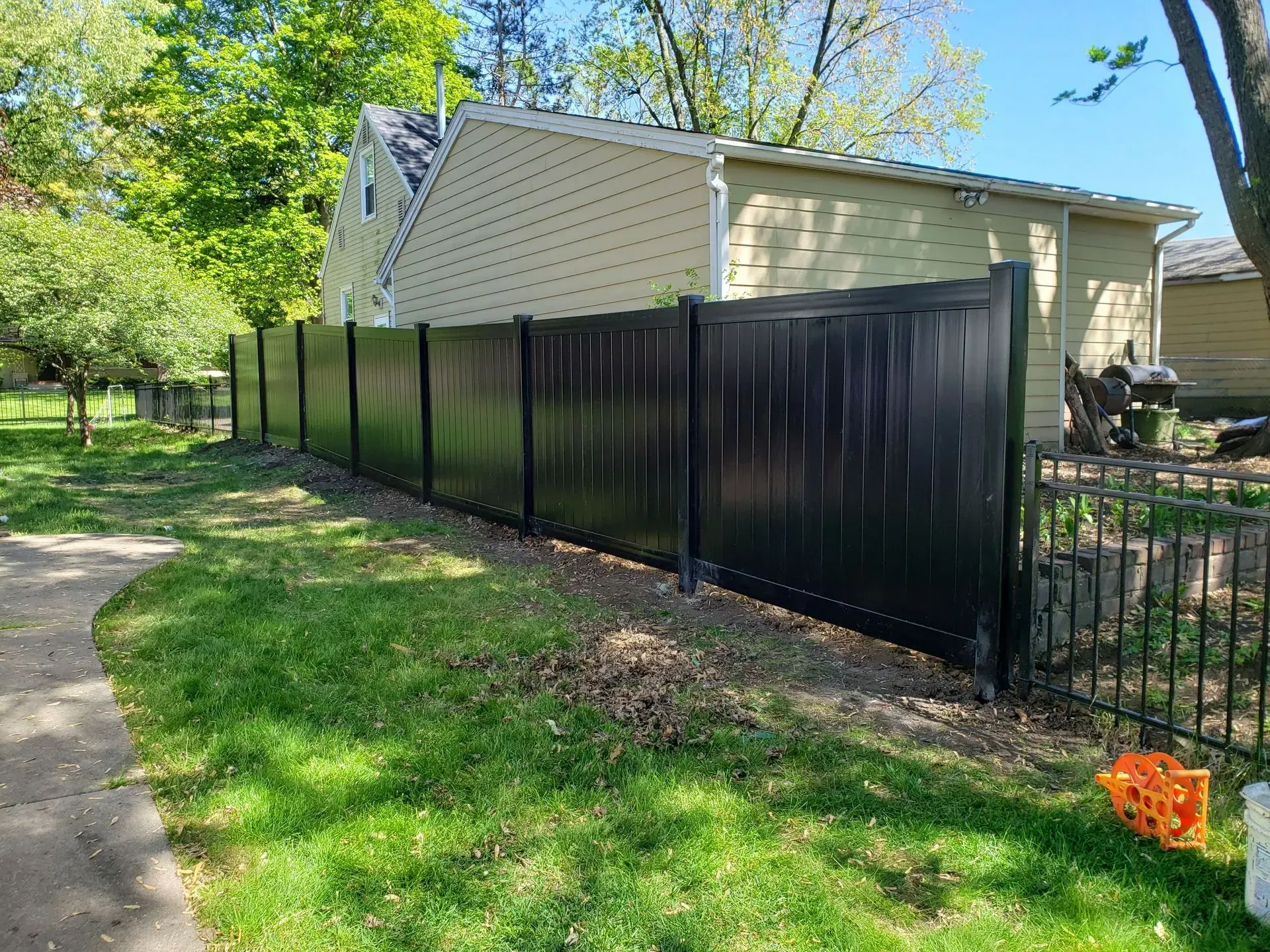 Black privacy fence along a yard next to a tan house. Green grass and trees are nearby.