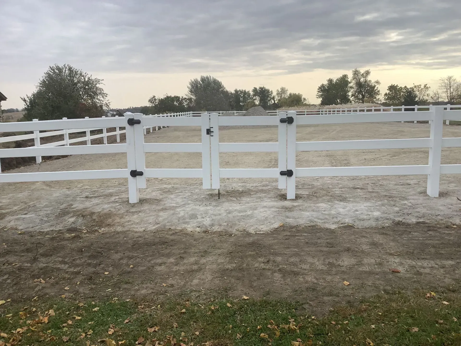 White horse fence and gate in a dirt arena, under a cloudy sky.