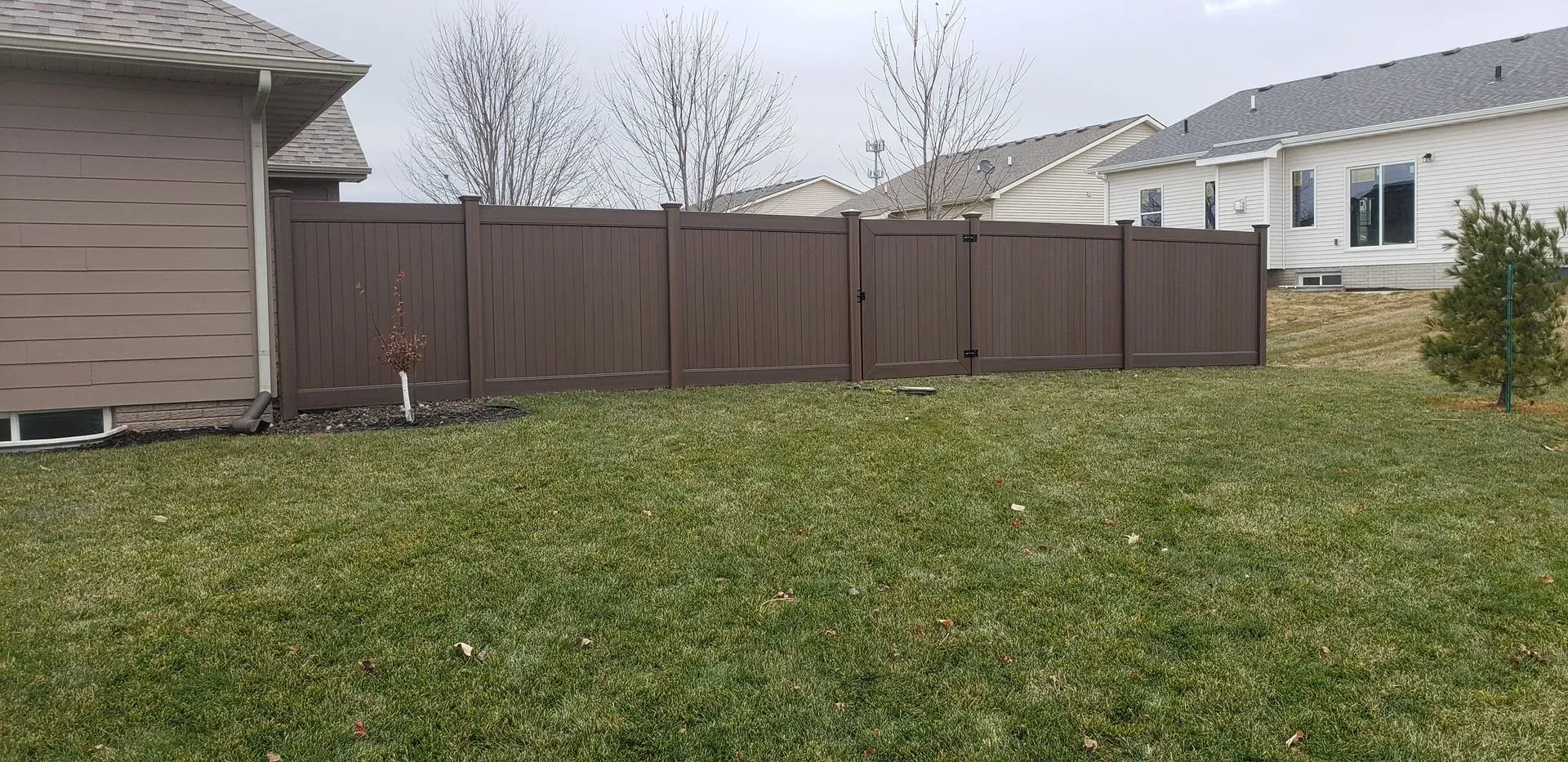 A brown fence borders a yard with green grass and a house under a cloudy sky.