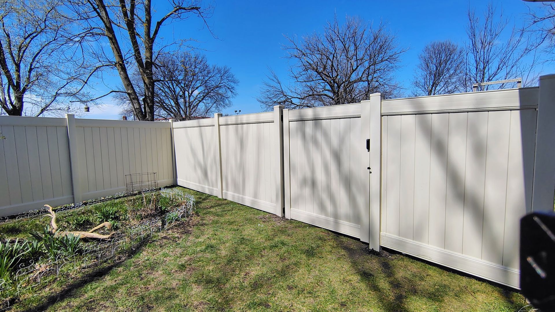 White vinyl fence along a grassy backyard under a clear blue sky, with bare trees in the background.