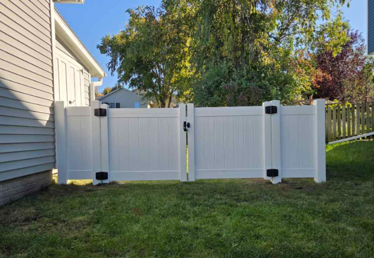 White vinyl fence panels beside a beige house in a grassy backyard with trees in the background