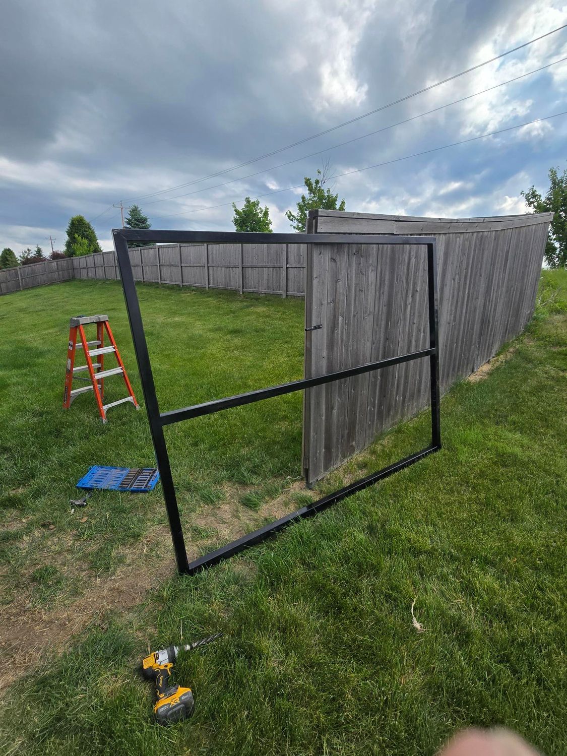 Black wooden gate panel leaning open in a grassy yard beside a fence, with a ladder nearby.