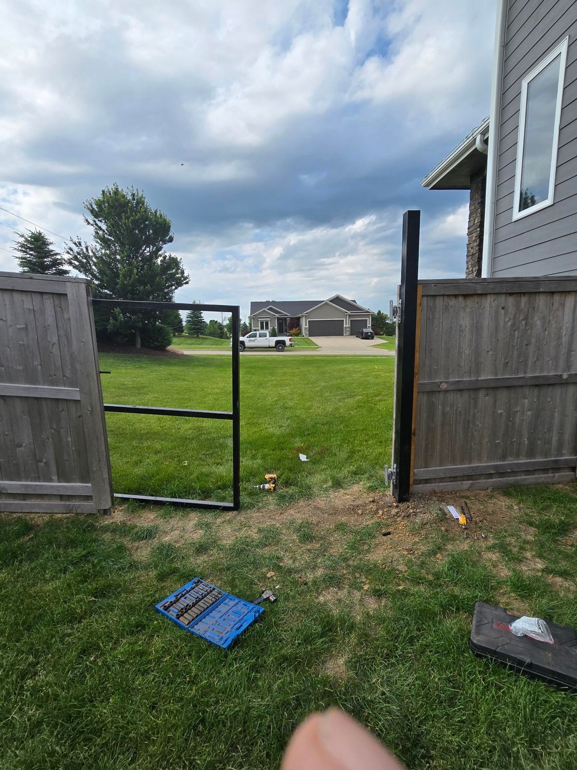 Backyard gate opening to a grassy yard and distant house, with blue panels on the ground and a gray house wall.