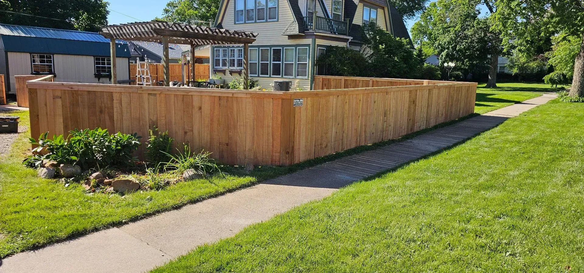 A wooden fence surrounds a yard with a sidewalk and green grass. A house is in the background.