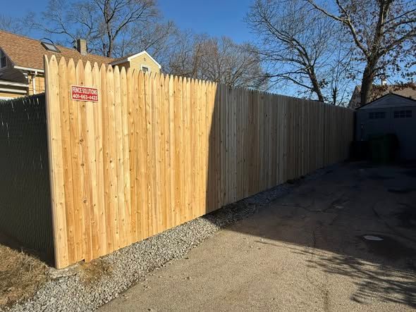 Wooden fence with pointed top along a paved area; sunny day.
