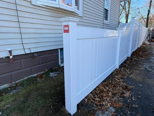 White vinyl fence along a house, with leaves on ground and a red sign on a post.