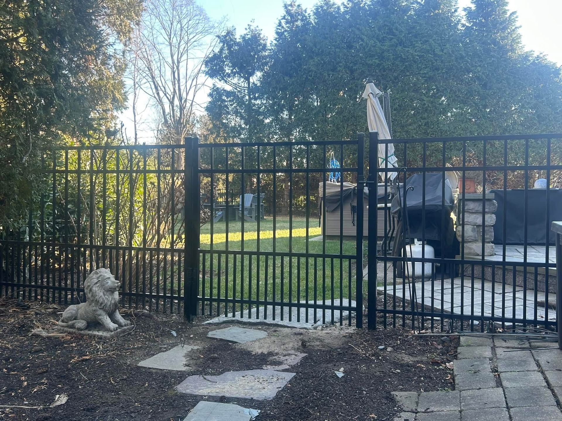Black metal fence surrounding a yard with a lion statue and stone patio. Trees in the background.