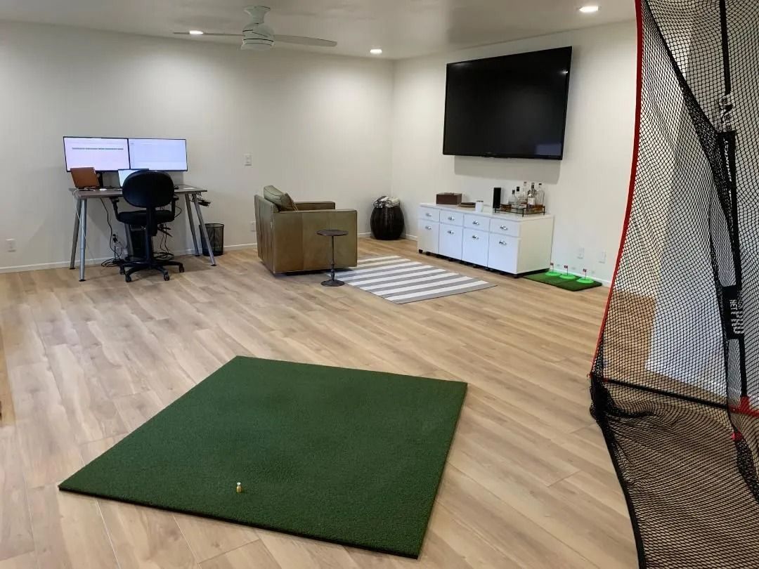 A home office/golf simulator setup with a golf mat, net, desk with monitors, couch, and TV.