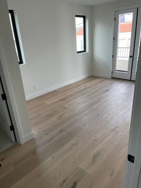 Empty room with light wood floors, white walls, black window frames, and a door to a balcony.
