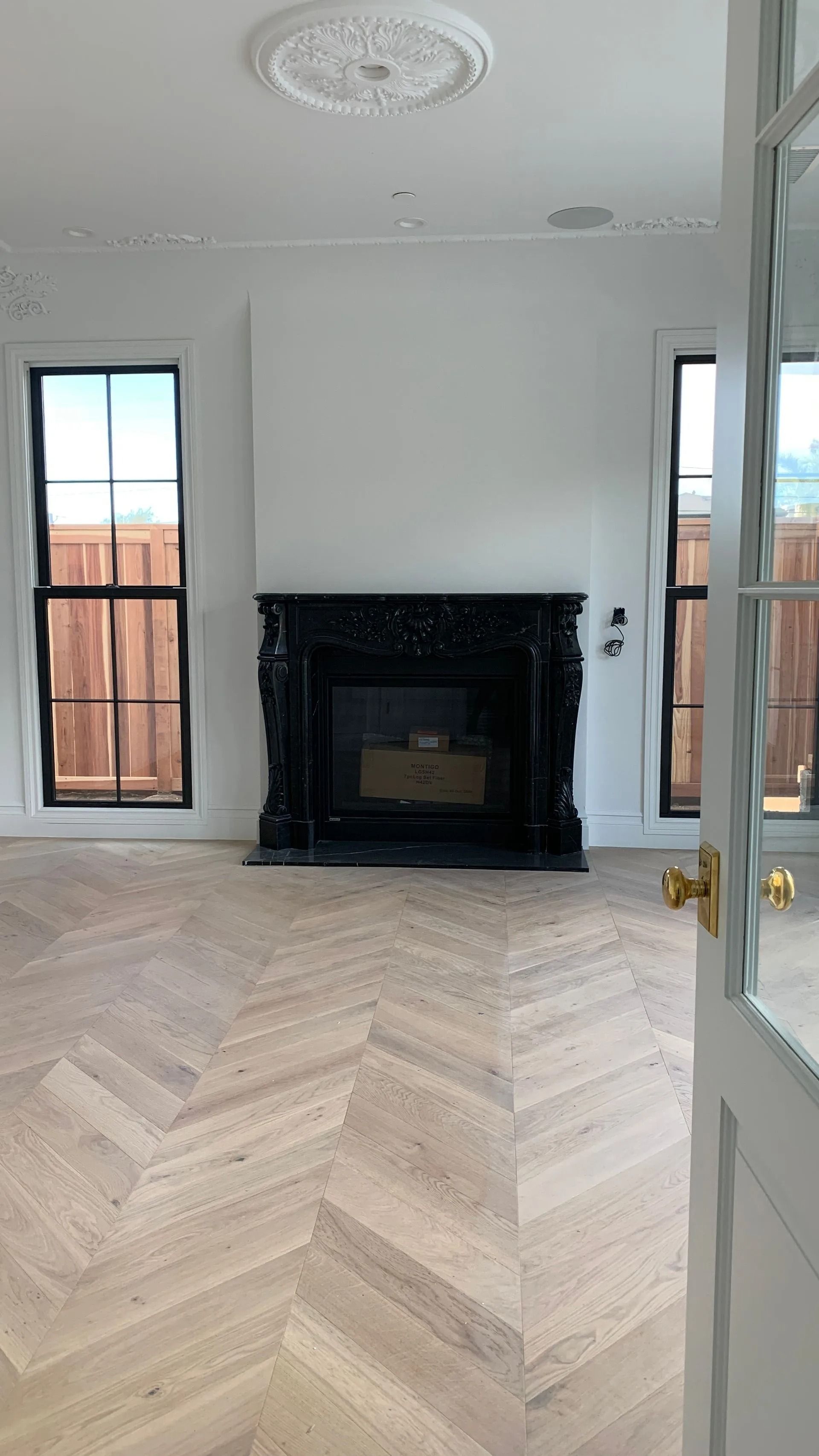 Empty room with herringbone wood floor, black fireplace, two windows, and ornate ceiling detail.
