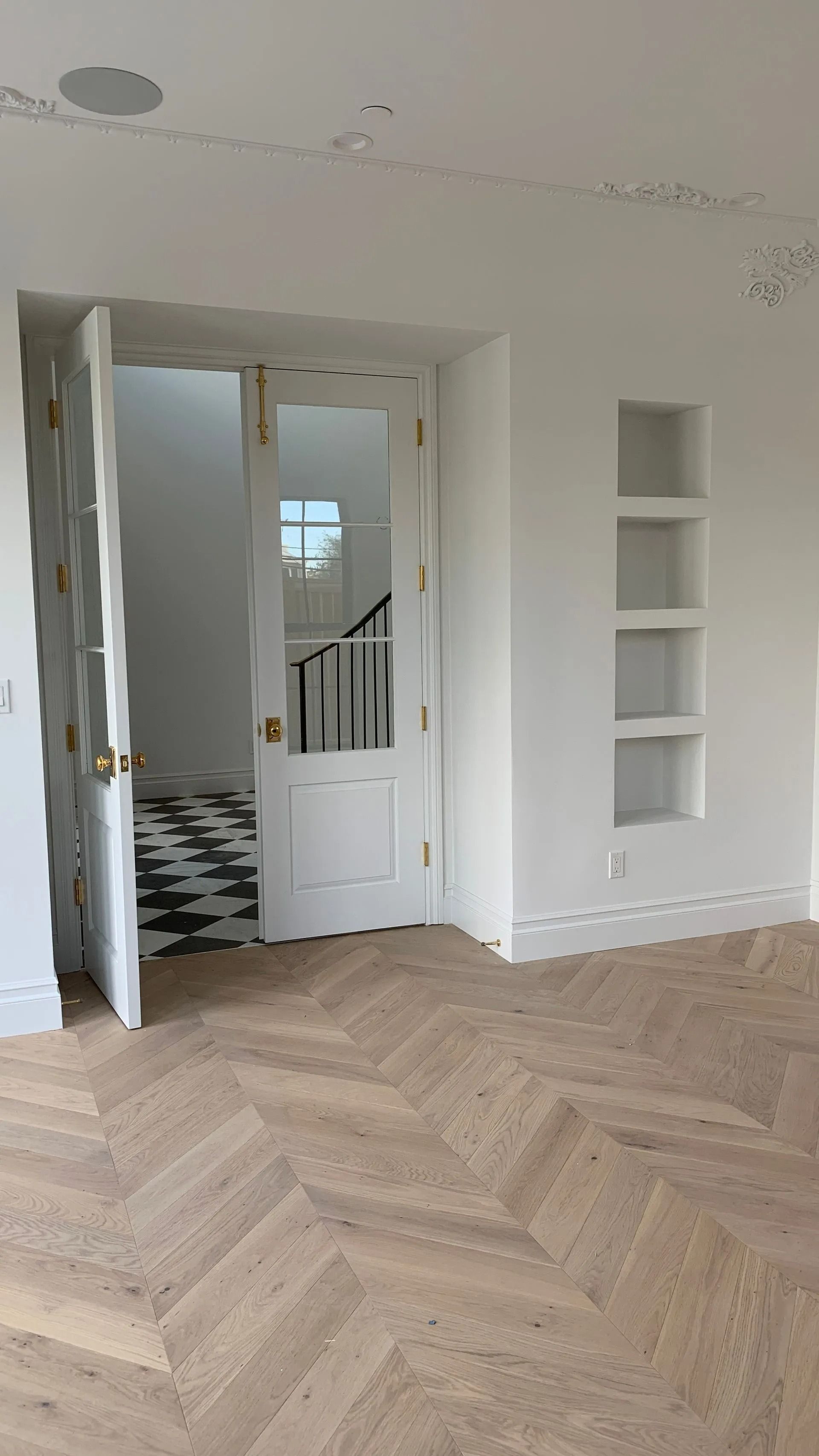 White room with parquet floor, doorway with stairs. Built-in shelves, black and white tiled floor in the doorway.