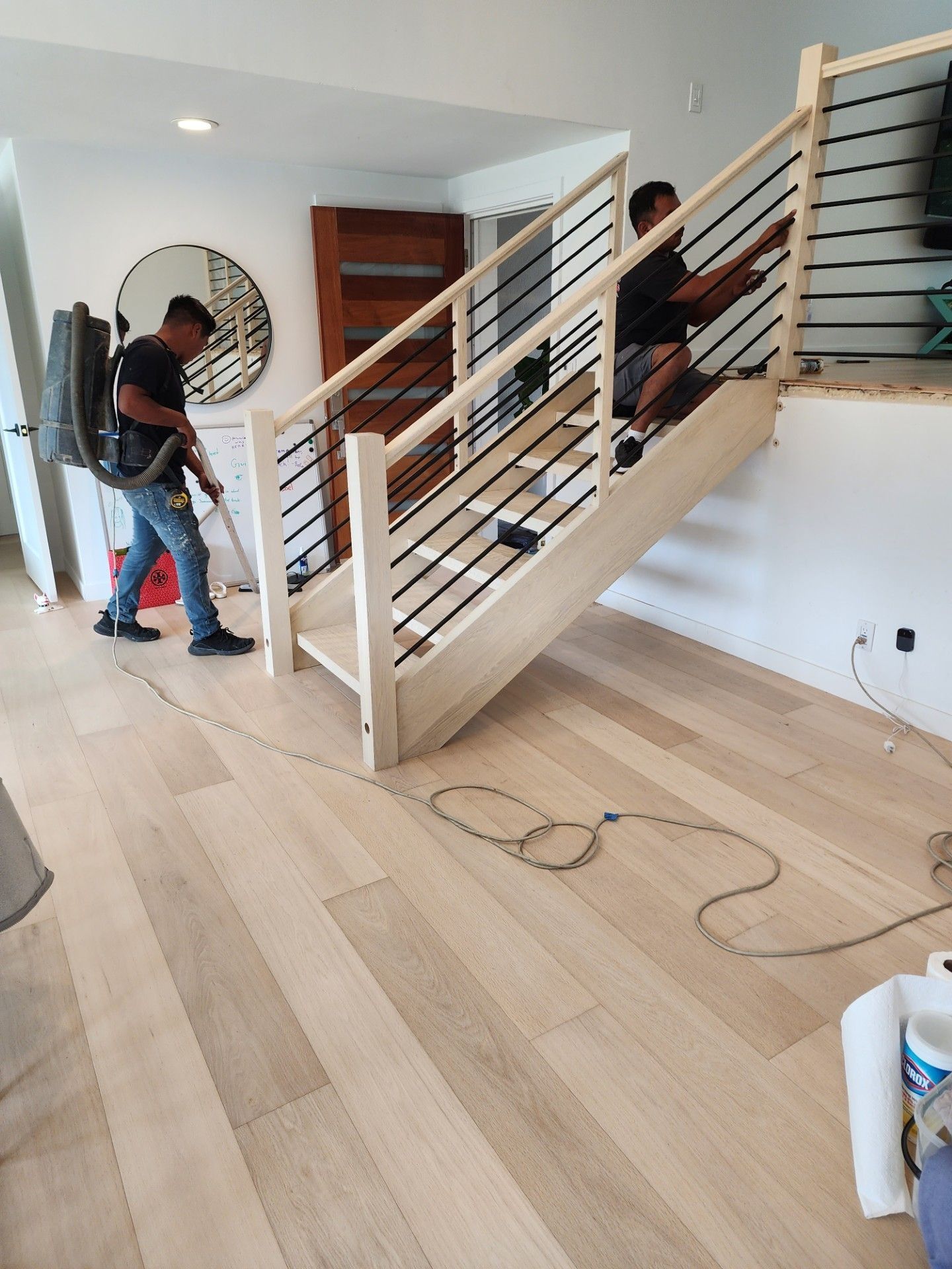 Two men cleaning a wooden staircase. One vacuums, the other adjusts the railing. Light wood floor.