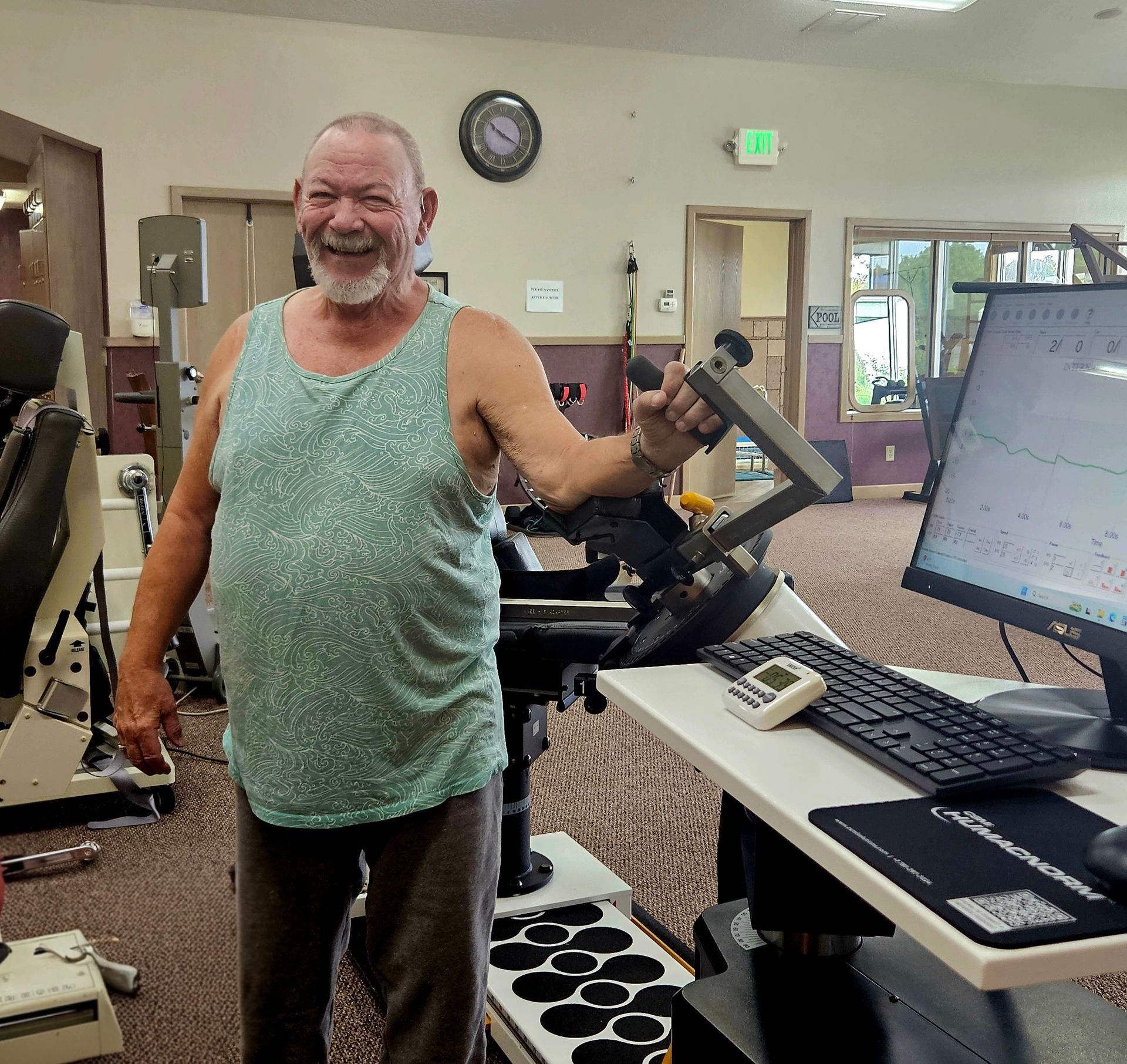 Smiling man in a gym, holding a cable machine handle beside a computer station.