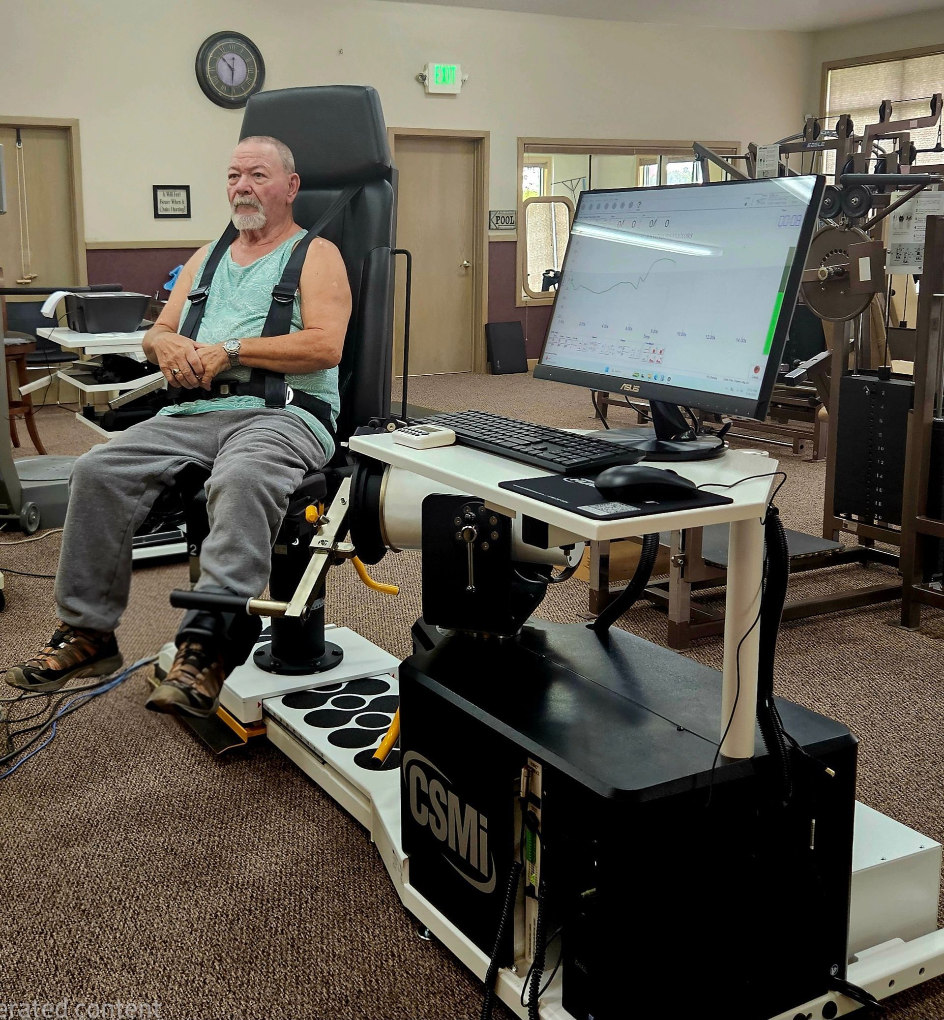Man seated in a motorized chair beside a large control console in a room with patterned carpet and a wall clock