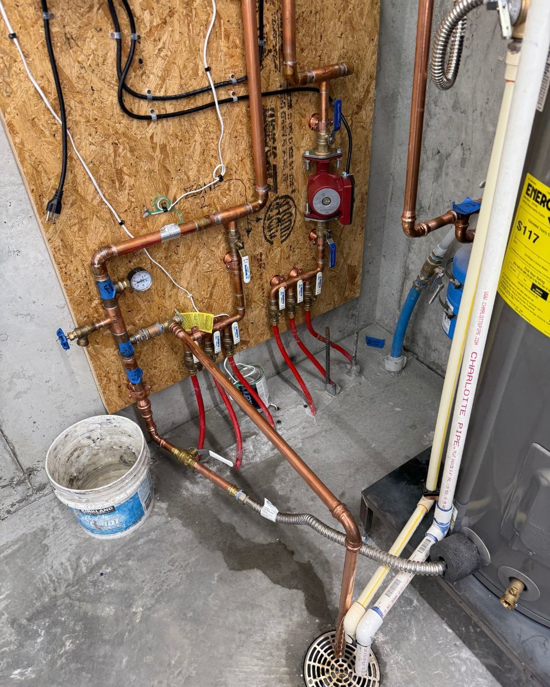 Plumbing wall with exposed copper pipes, valves, red hoses, and a bucket on a concrete floor.