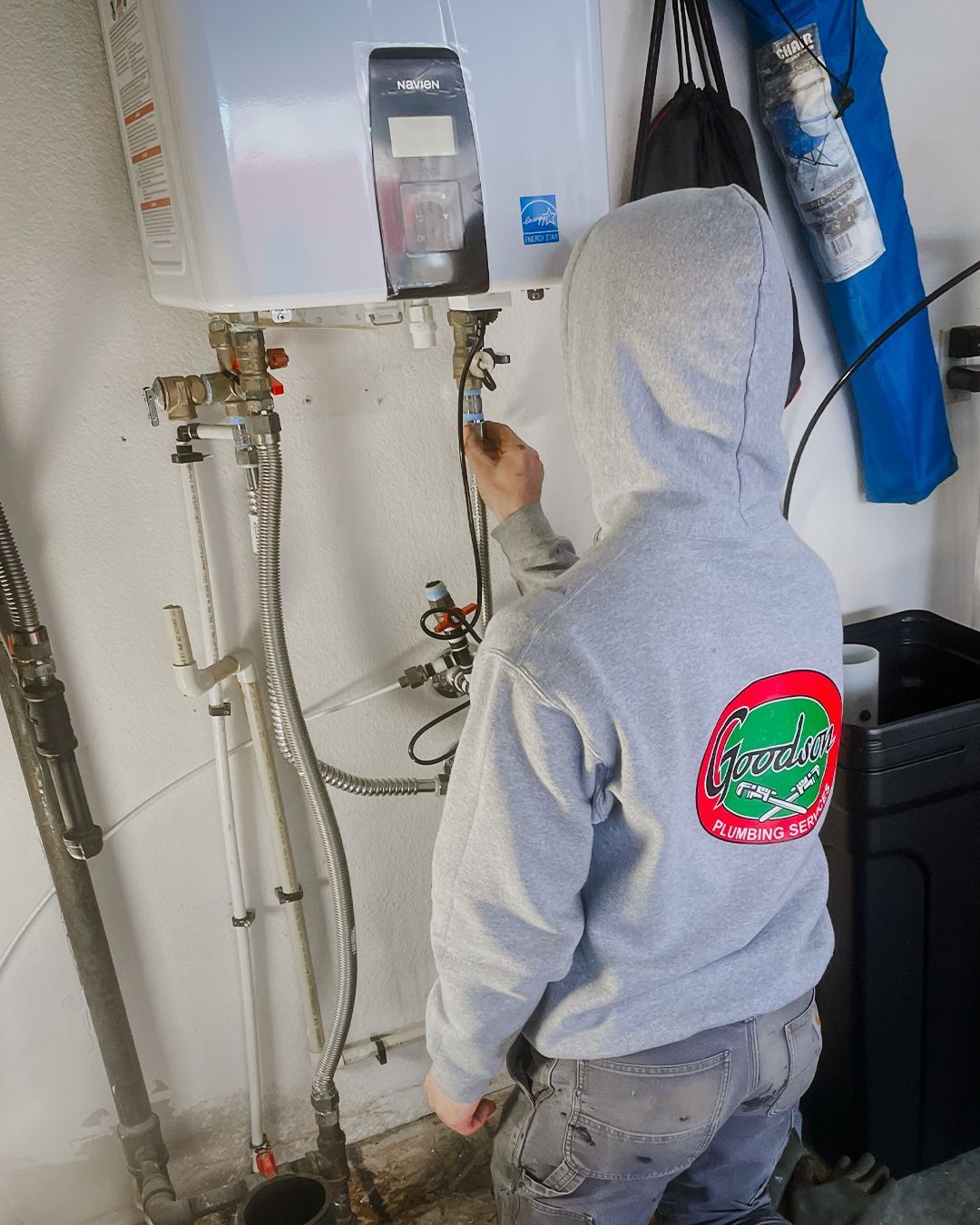 Person in a gray hoodie inspects plumbing equipment in a utility room.