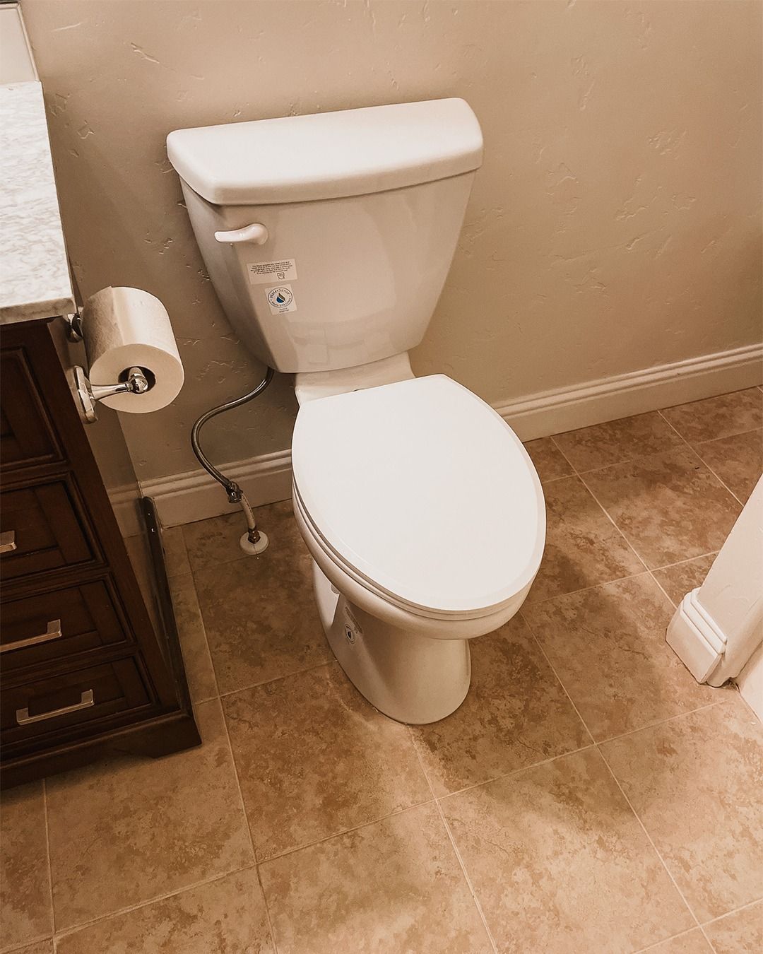 White toilet in a beige-tiled bathroom with a toilet paper roll on the wall and a dark wood cabinet nearby.