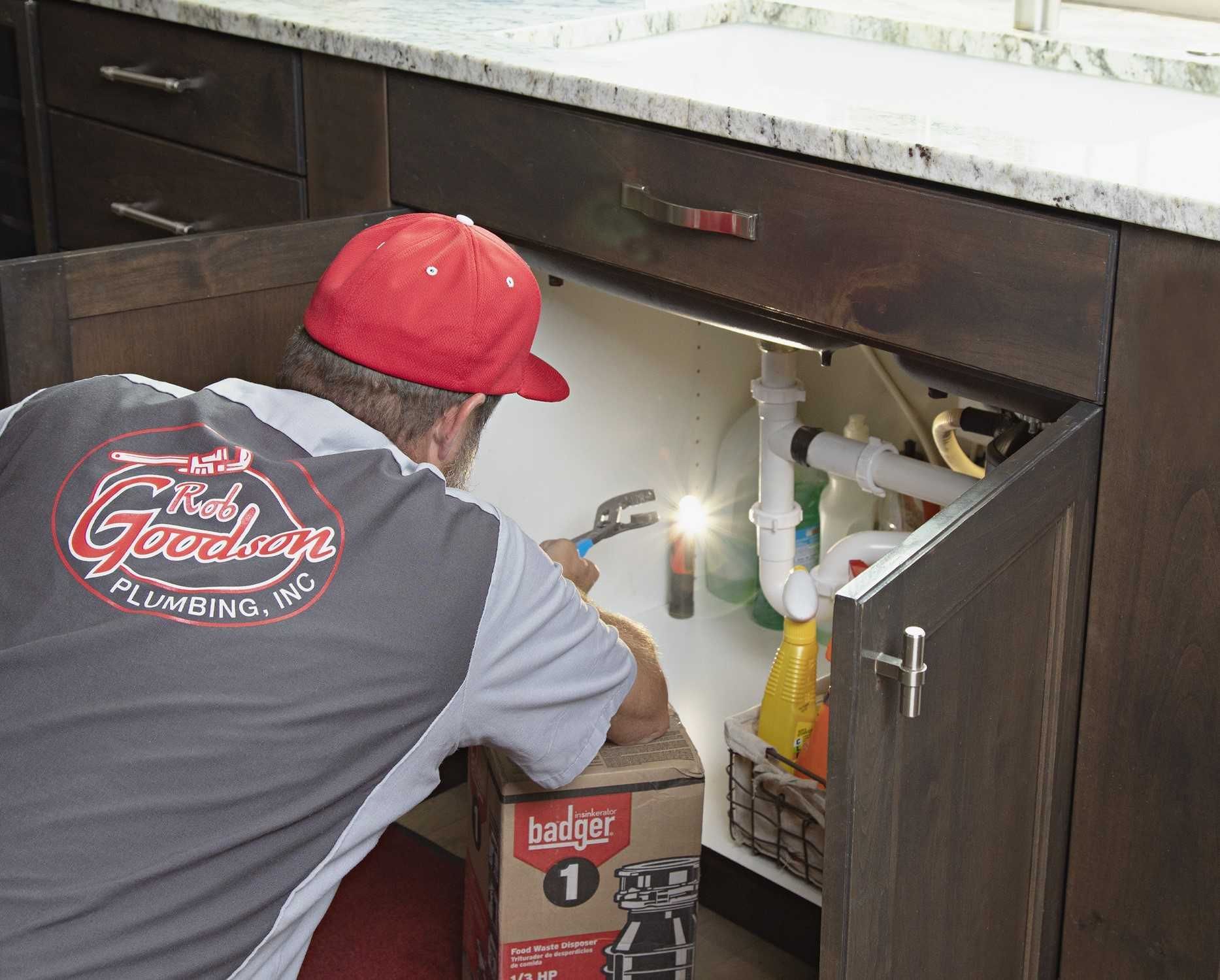 Worker inspecting under-sink pipes in a kitchen cabinet with a flashlight.