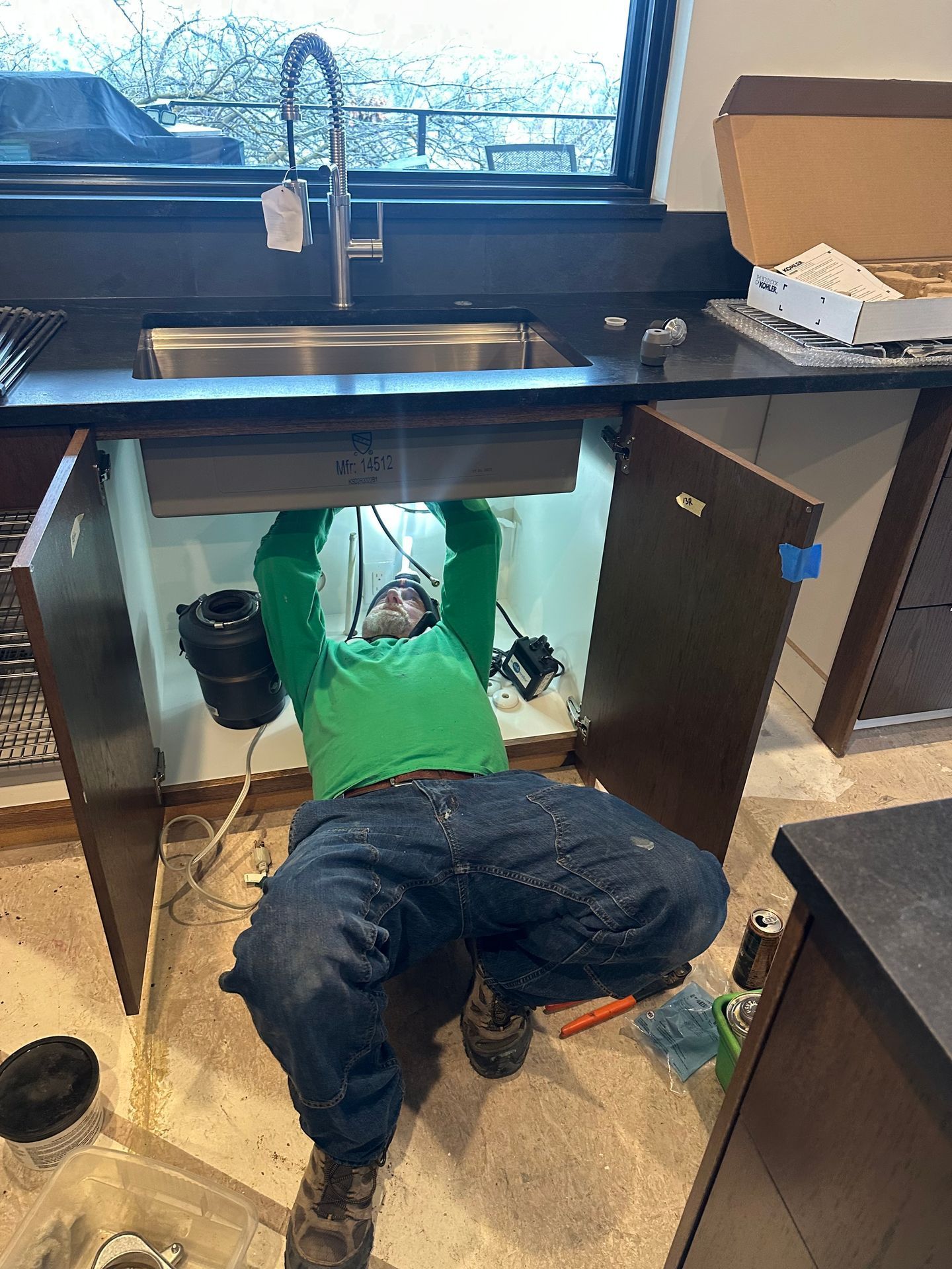Person lying under a kitchen sink cabinet, reaching up into the open sink area during plumbing work.
