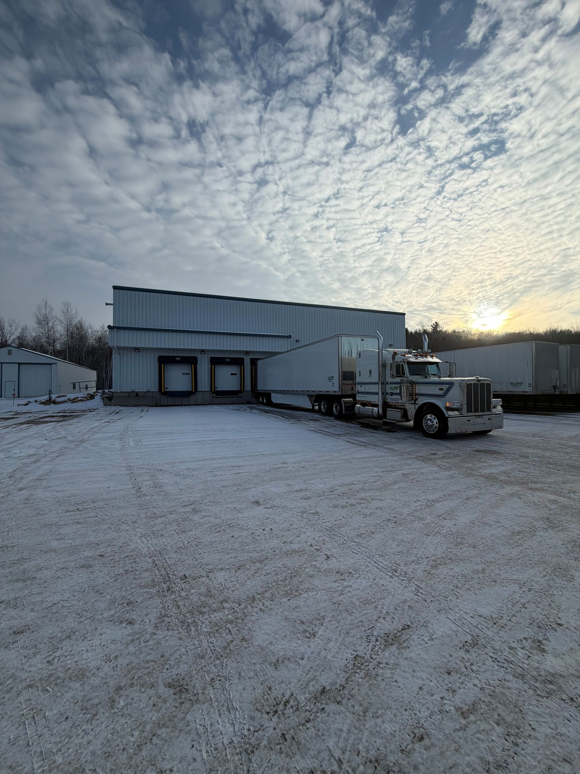 Semi-truck at loading docks of a white industrial building in a snowy lot under a cloudy sky at sunset.