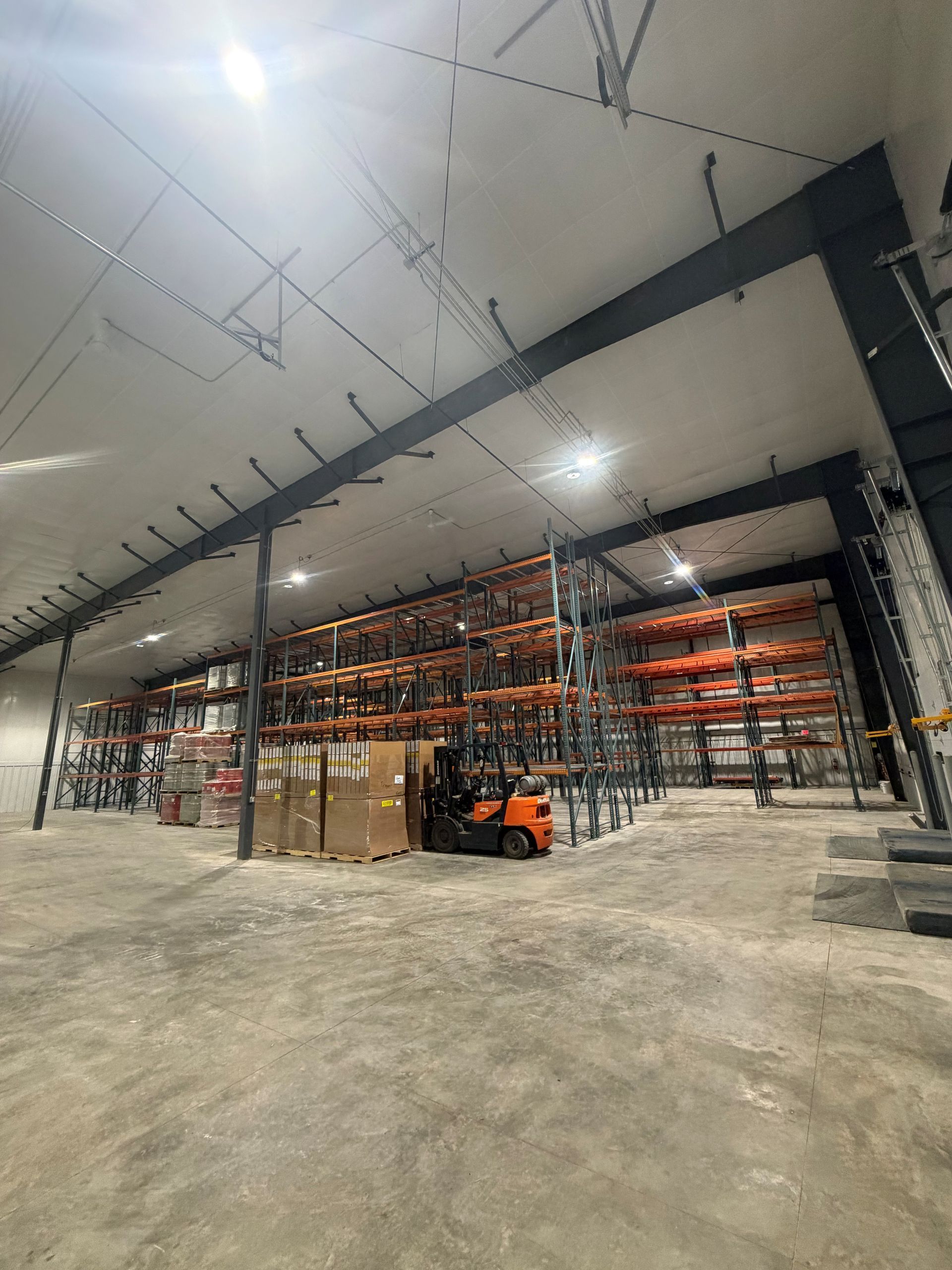 Warehouse interior with forklifts, pallets, and shelving units. Grey concrete floor, black metal beams, and fluorescent lights.