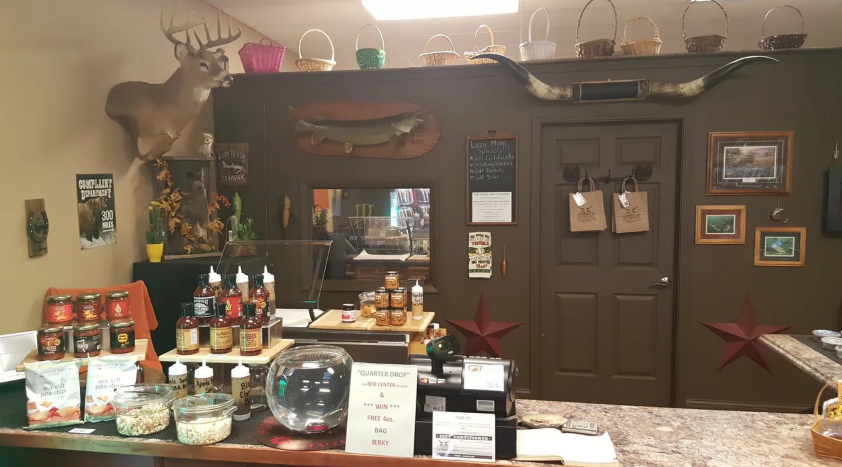 Interior of a shop with a display of jars, sauces, and baskets. Taxidermy head and horns decorate the wall.