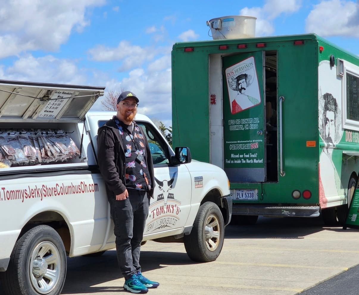 Man stands by white pickup truck and green food truck; sunny day.
