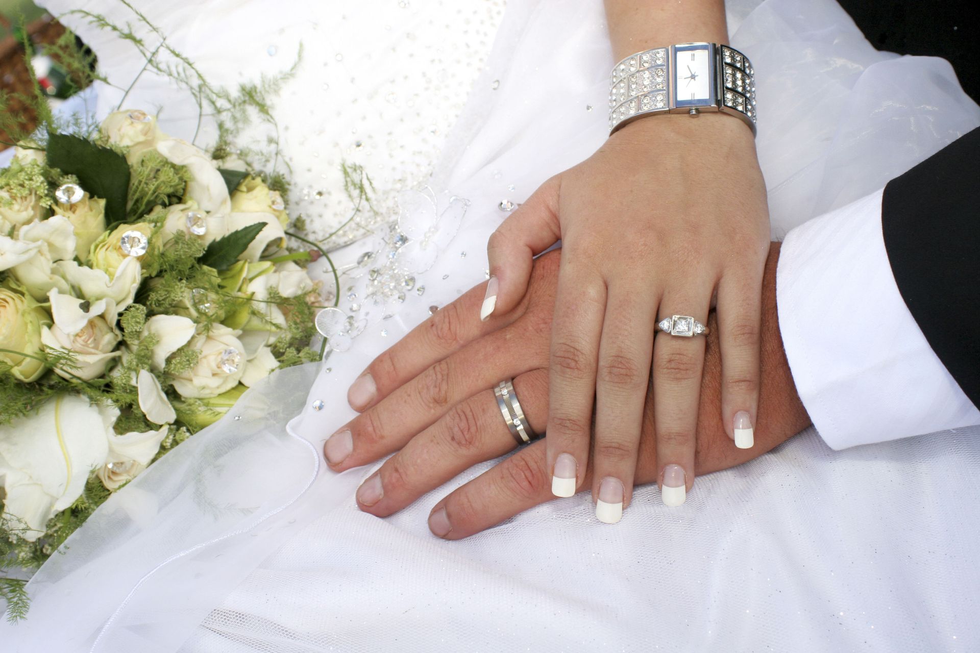 Hands with wedding rings resting on a white dress beside a bouquet of white flowers