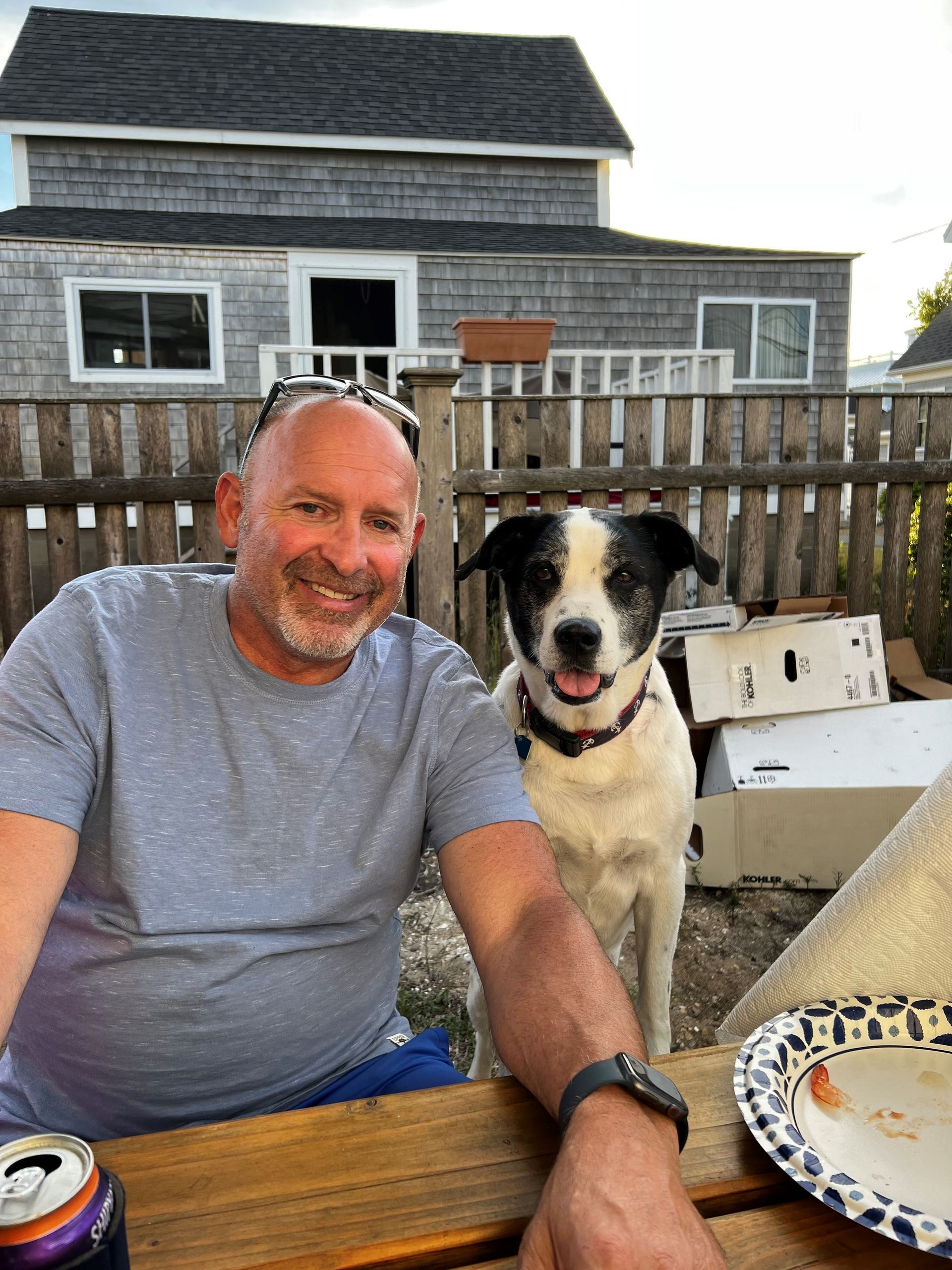 Man smiles next to a dog outside, near a house with a wooden fence. The dog has black and white fur.