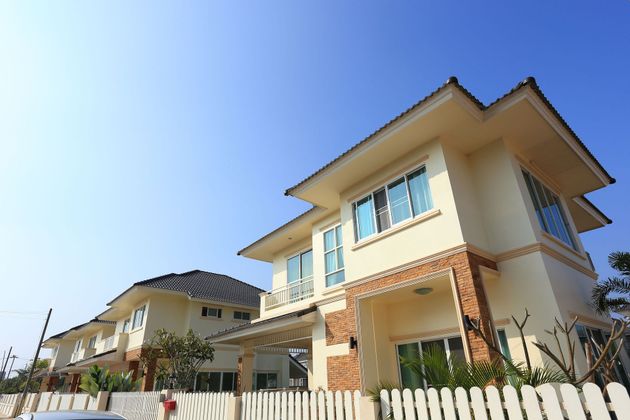 Two-story beige house with stone accents and a white fence under a clear blue sky