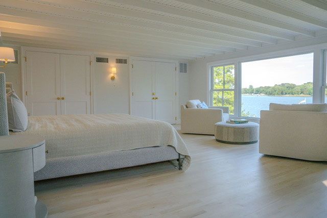 Bright white bedroom with large windows, a bed, two chairs, and a lake view