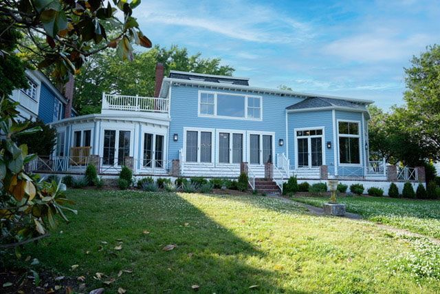 Blue house with white trim and large windows, set behind a green lawn and trees on a sunny day