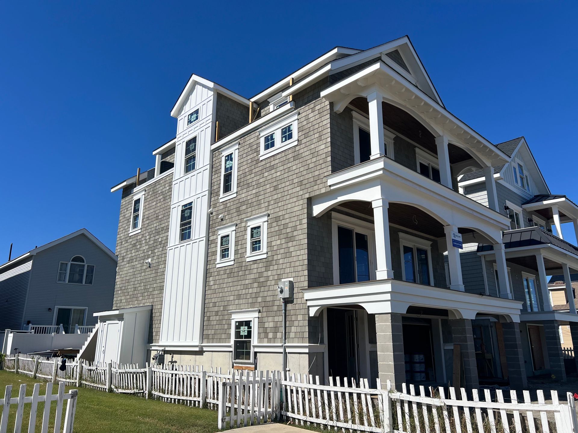 Large gray house with white trim and balconies on a sunny street behind a white fence